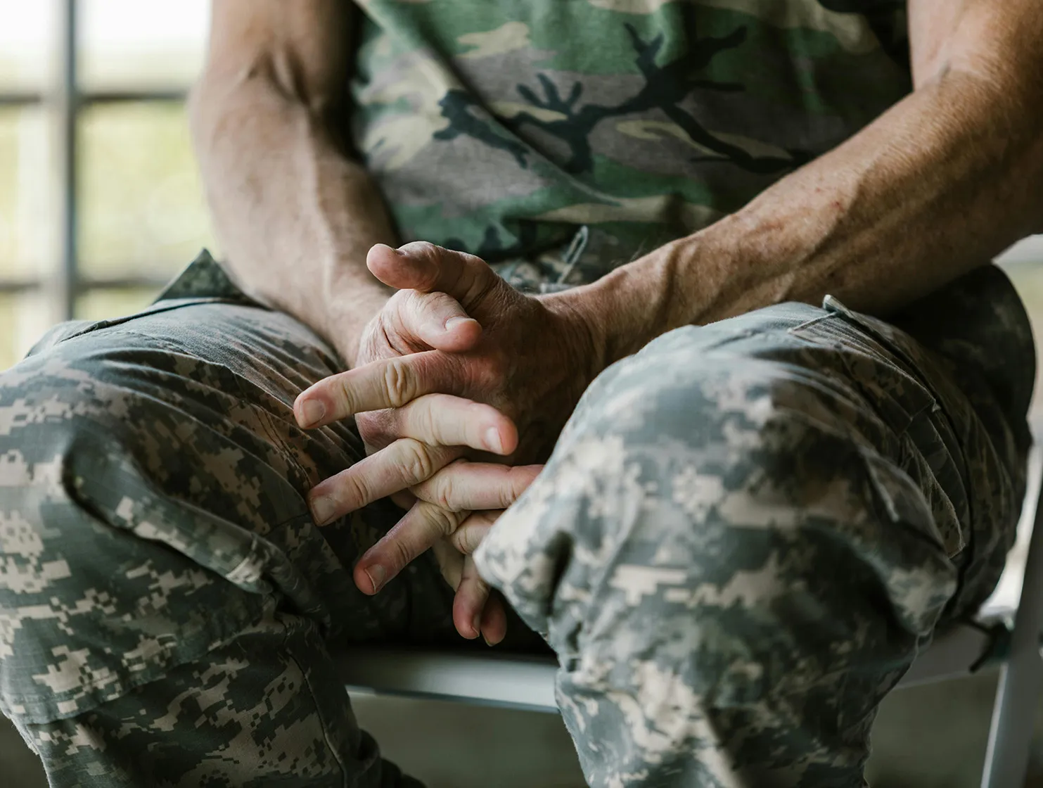 Close-up of a person wearing camouflage military uniform sitting with hands clasped between their knees.