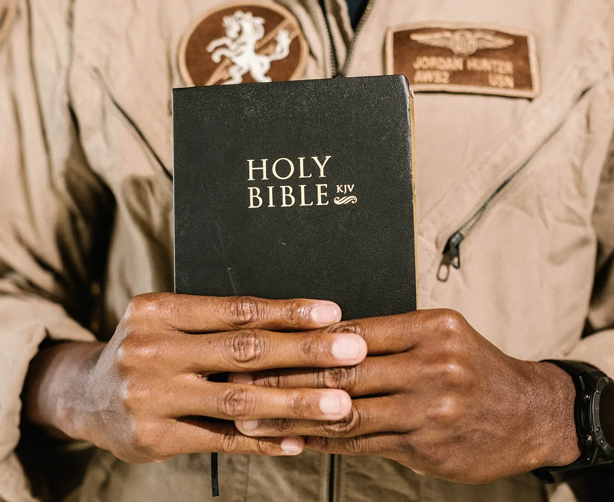 Person wearing a beige jacket holding a black Holy Bible KJV with both hands.