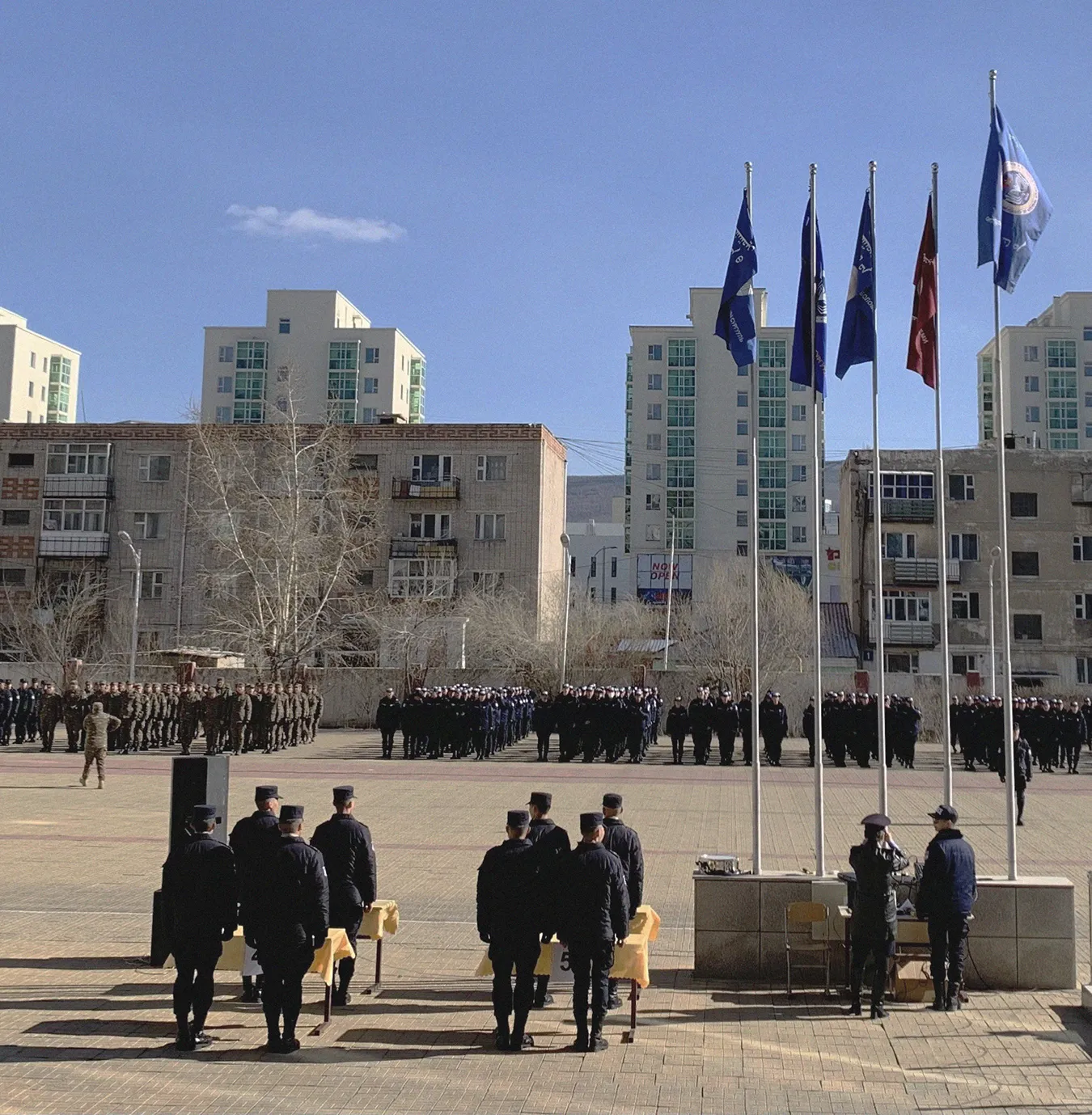 Uniformed personnel standing in formation outdoors near flagpoles with flags, in front of mid-rise buildings under a clear blue sky.