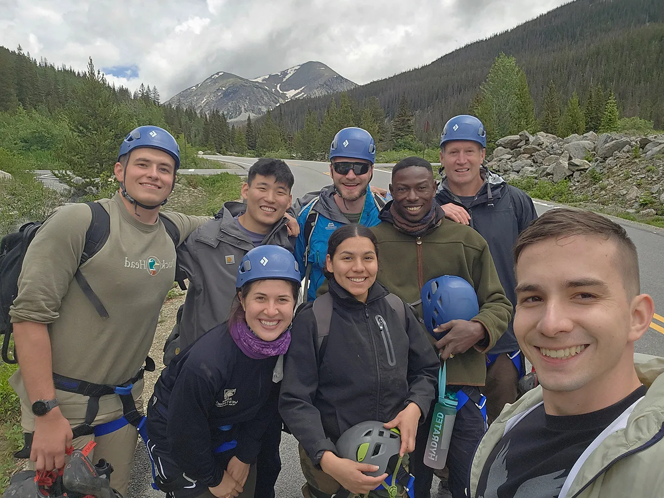 Group of seven smiling climbers in helmets posing for a selfie on a mountain road with forest and snowy peaks in the background.