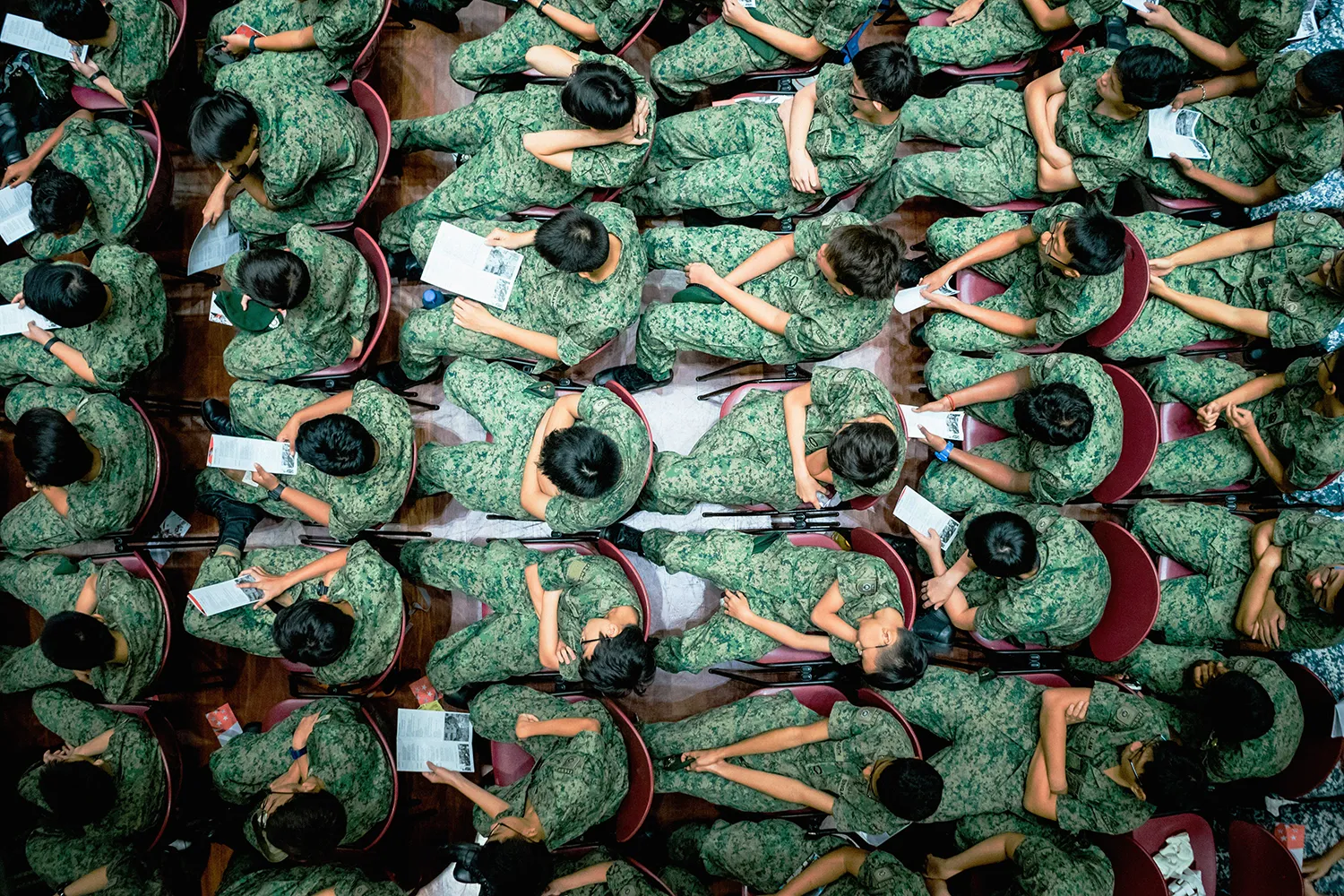Overhead view of soldiers in green camouflage uniforms seated closely in rows holding papers during a briefing or class.