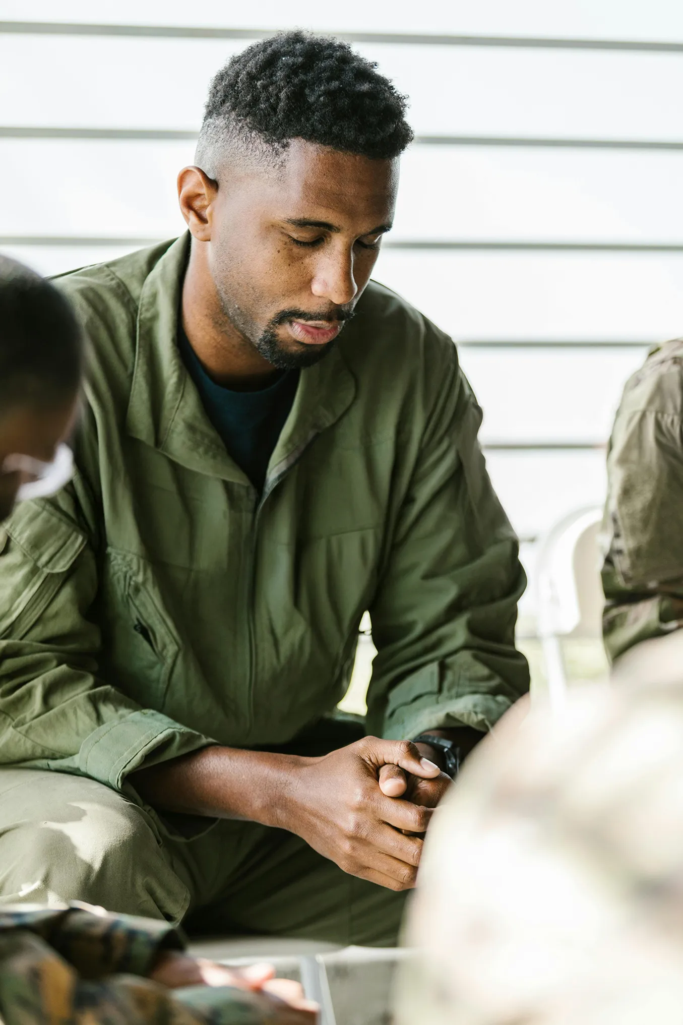 Soldier in green military uniform sitting with hands clasped and head bowed in a moment of reflection or prayer.