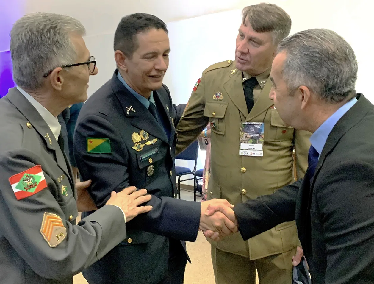 Four men, three in military uniforms and one in a suit, shaking hands and conversing indoors.