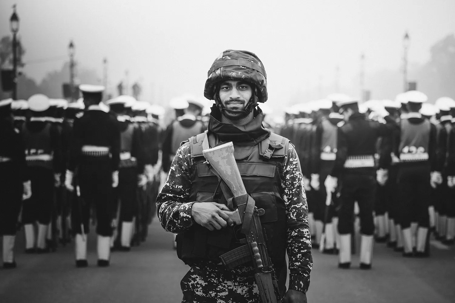 Smiling soldier in camouflage uniform and helmet holding a rifle with a formation of uniformed soldiers marching in the background.