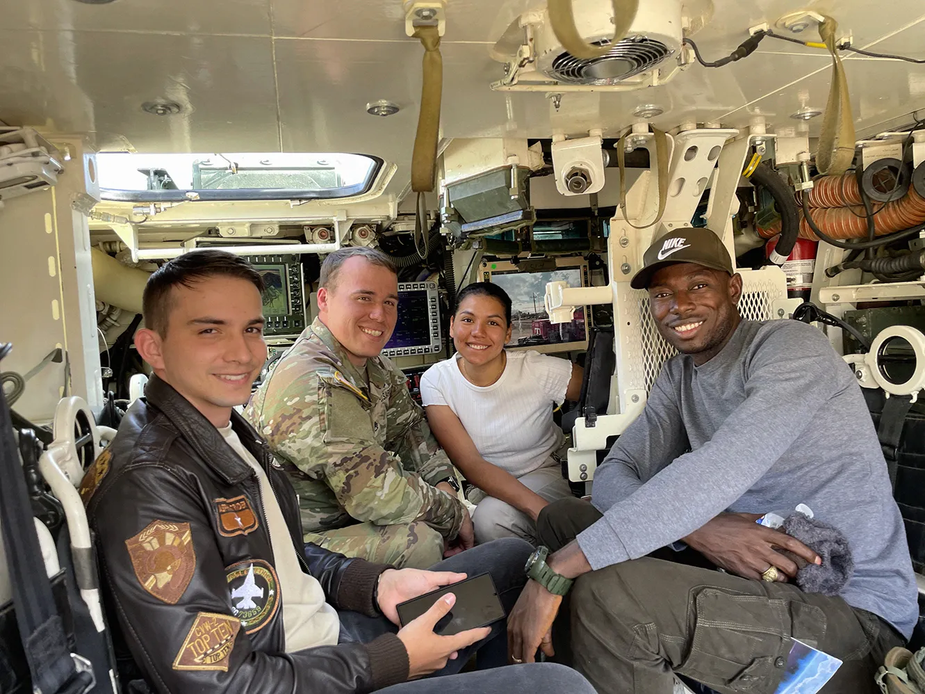 Four people sitting inside a military vehicle, smiling at the camera; two men wear casual clothing, one man is in military uniform, and a woman wears a white shirt.